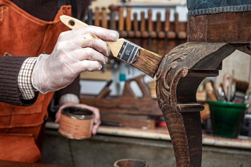 Close-up of a carpenter restoring an antique chair with wood wax and brush in a furniture workshop.